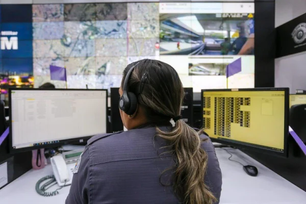 Imagem colorida mostra mulher de costas, com cabelos lisos e longos presos em um rabo. Ela está em frente a duas telas de computador que funcionam como central telefônica do 190 - Metrópoles