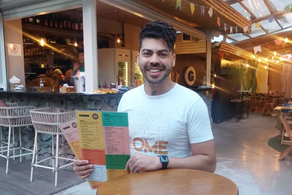 Chef Rafael Silva, sentado à mesa, segurando cardápios físicos