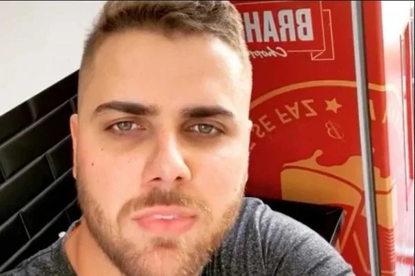 Na imagem colorida, um homem está posicionado à esquerda. Ele usa roupa cinza e olha seriamente para acamera