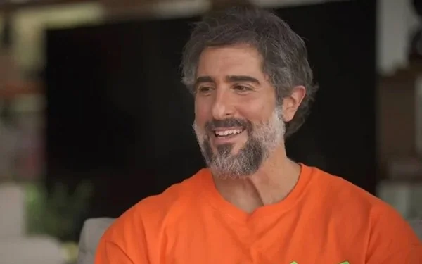 Na imagem colorida, um homem está posicionado no centro. Ele tem cabelos curtos e grisalhos e usa camiseta laranjada