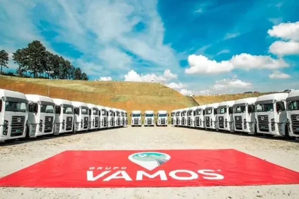 Imagem da frota de caminhões do Grupo Vamos, um ao lado do outro. Ao centro, o logotipo da empresa em um cartaz vermelho sobre o chão. Ao fundo, árvores e céu azul - Metrópoles