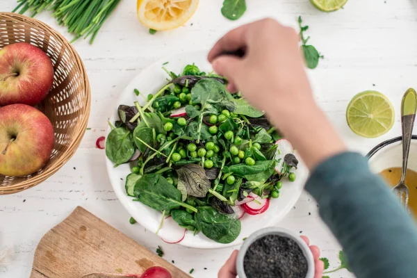 Foto colorida de prato branco com verduras e legumes, enquanto mulher coloca tempero na salada - Metrópoles