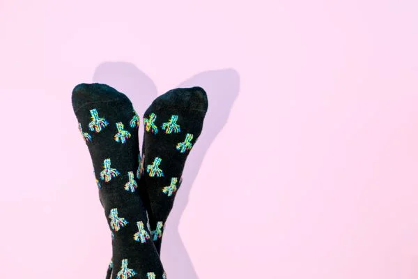 Pés cruzados para cima usam meias divertidas pretas e enfeitadas com detalhes coloridos. O fundo é rosa - Metrópoles