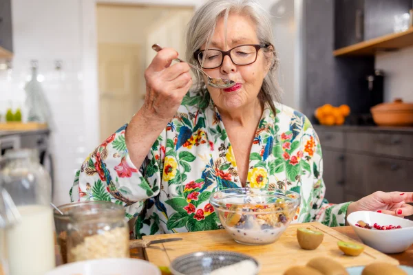 Foto colorida de mulher mais velha de alimentando sentada à mesa - Crononutrição: saiba como o horário em que comemos influencia na saúde - Metrópoles