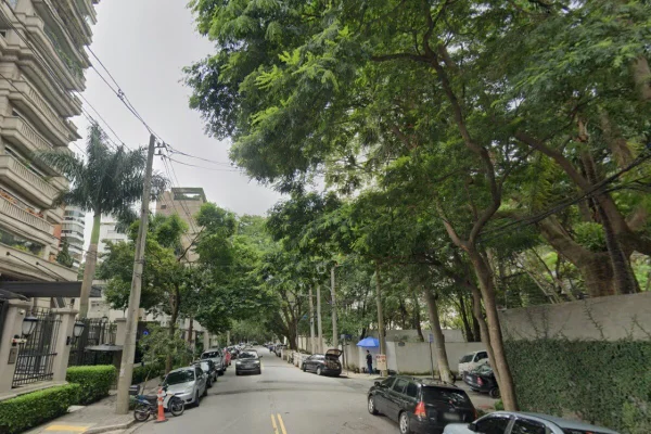 Imagem colorida de uma rua com um prédio ao lado esquerdo e uma árvore do lado direito, com carros estacionados
