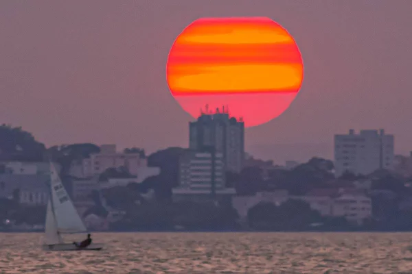 Na foto vemos uma pessoa velejando uma cidade e o sol ao fundo