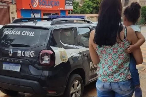 Imagem colorida: mulher carrega criança de 2 anos resgatada de sequestro no colo, ambas de costas, em frente à viatura da polícia - Metrópoles