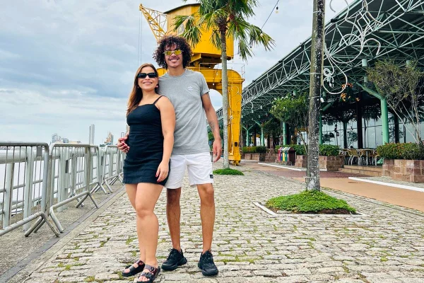Mulher com roupa preta e homem com roupa cinza em frente a um rio