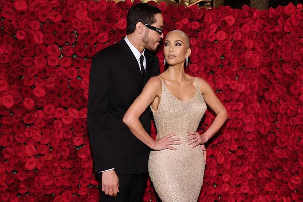 Empresária Kim Kardashian e o namorado, o comediante Pete Davidson, no evento MET Gala em 2022. Os dois posam para foto abraçados em um fundo vermelho de rosas de verdade. Ele usa um terno clássico preto, com camisa branca e gravata também preta. Kim usa um vestido rente ao corpo, semi transparente e com pequenas contas de cristais. A peça foi usada pela atriz Marilyn Monroe nos anos 1960.