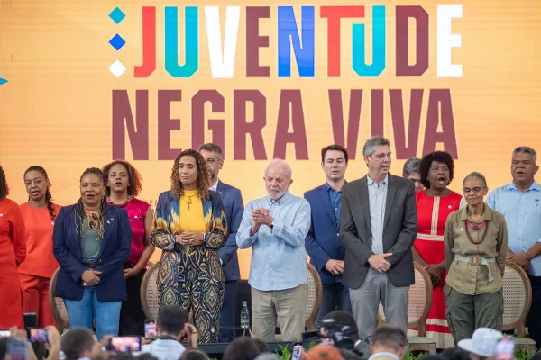 Imagem colorida de evento com frase "Juventude Negra Viva" escrita colorida em telão. Na frente, presidente Lula e ministros - Metrópoles