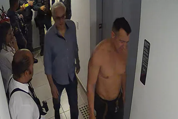 Homem branco de cabeo branco e camisa social junto com homem branco, sem camisa, em corredor de prédio - Metrópoles