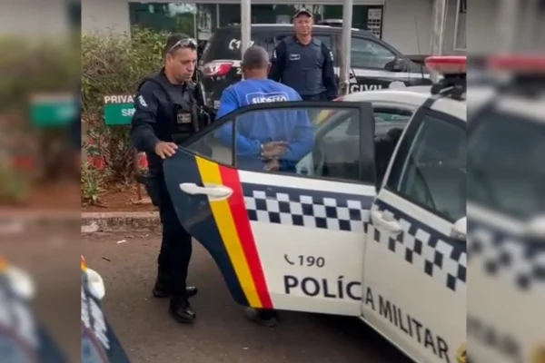Imagem de um homem de camiseta azul descendo de uma viatura da PMDF em direção à delegacia, acompanhado por dois policiais
