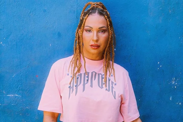 A cantora Valesca Popozuda posa para foto sob parede azul. Ela olha para a câmera, série, usa tranças com miçangas e blusa rosa com roxo - Metrópoles