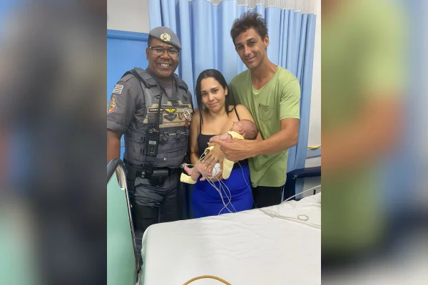 Imagem colorida mostra bebê no colo dos pais ao lado de um policial militar (PM) - Metrópoles