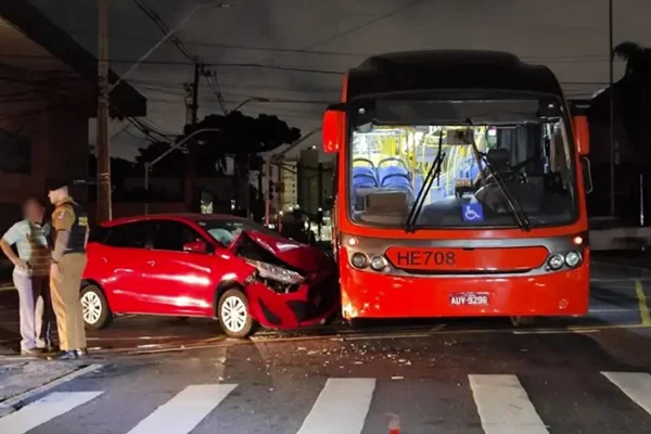Foto de acidente entre carro e ônibus biarticulado em Curitiba. Policial e homem estão ao lado - Metrópoles