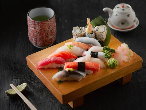 Tabuleiro com peças de sushi e outros tipos de comida japonessa com hashis ao lado e copo de chá verde - Metrópoles