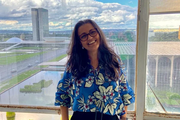 Foto colorida de mulher de blusa azul em frente a um vidro coom vista para o congresso Nacional - Metrópoles.