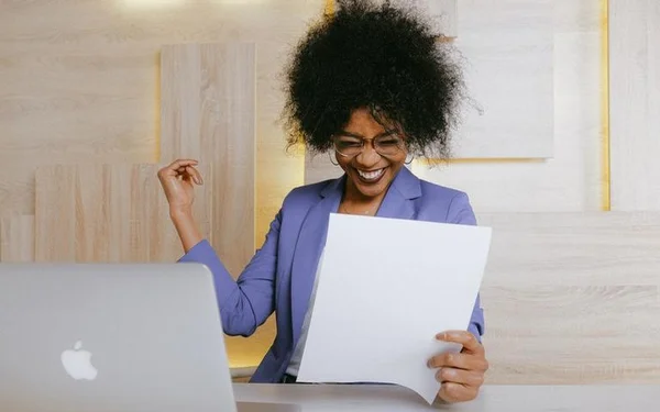 Mulher com papel na mão em frente a computador com sorriso de felicidade