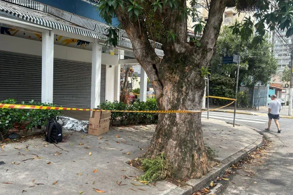 Imagem colorida mostra área de calçada isolada com fitas coloridas em laranja e vermelho. Dentro da área isolada, é possível ver uma mochila preta no chão e, ao lado, um corpo deitado, coberto por manta de alumínio - Metrópoles