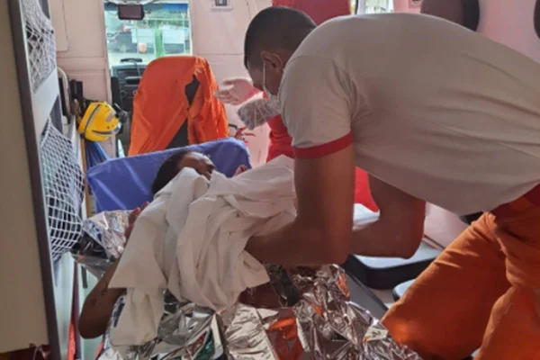 na foto temos o interior de uma ambulancia com uma mulher deitada coberta por uma manta de aluminio e um homem de camiseta branca entregando um bebe enrolado em um pano branco