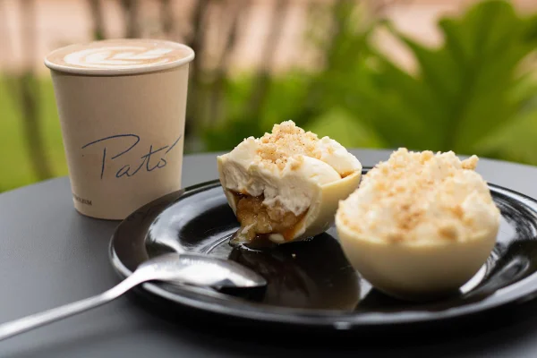 na foto vemos um prato preto com ovo de colher branco e outro cortado ao meio onde se ve banana caramelada e chantilly e um copo de papel com café
