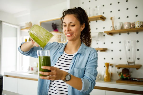 Foto colorida de mulher segurando liquidificador e colocando suco em copo - Metrópoles