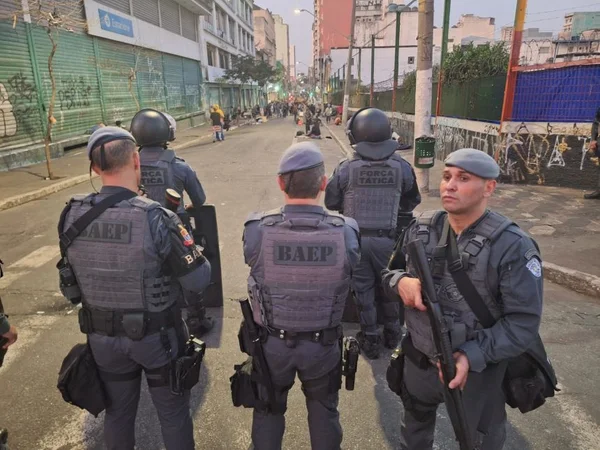 Foto colorida de cinco policiais militares armados em operação na cracolândia com usuários de drogas ao fundo - metrópoles
