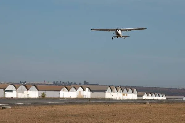 Fotografia colorida de avião chegando em pista de pouso com hangares ao fuundo