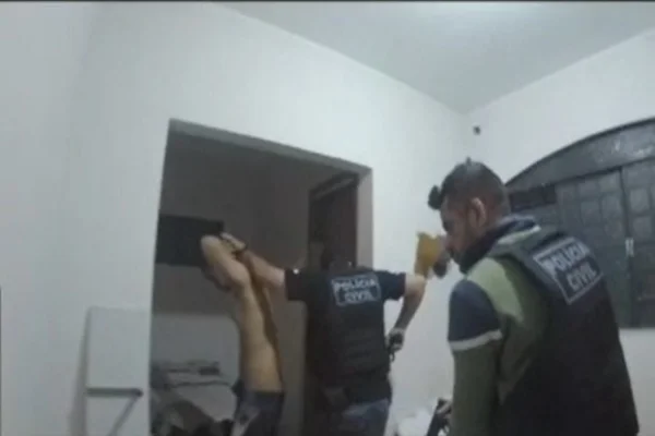 policiais e homem sem camisa com mãos na cabeça em sala de estar