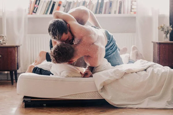 Foto colorida de dois homens sem camisa e de calça jeans se abraçando em ciam de um colchão no chão - Metrópoles