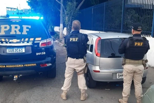 Imagem colorida mostra dois policiais de costas, com coletes com a sigla PRF. Eles estão voltados para a traseira de um veículo Corsa - Metrópoles