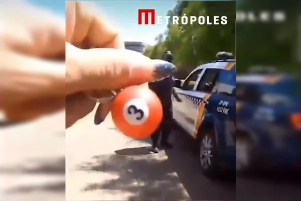 Imagem colorida de uma mulher segurando um chaveiro vermelho escrito o número 3. Ao fundo, um policial fardado encostando em uma viatura da PMDF