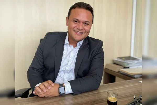 Homem de camisa social e blazer com braço em mesa de madeira e sorrindo