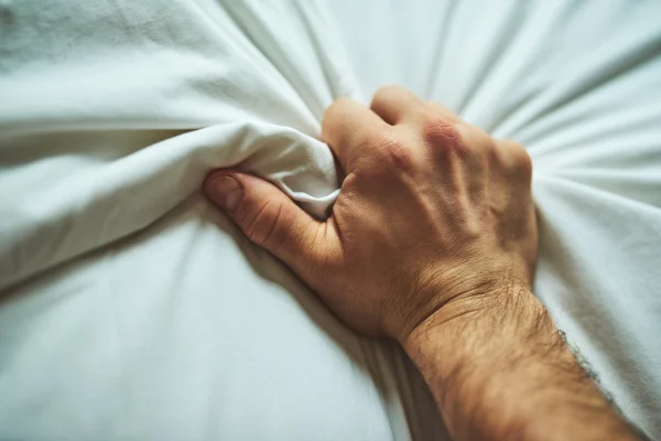 Foto em close da mão de um homem irreconhecível agarrando um lençol com prazer