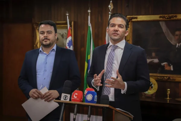 Imagem colorida mostra Rodrigo Goulart, homem pardo, de cabelo preto e barba preta aparada, vestindo terno escuro e camisa azul clara, e Rubinho Nunes, homem branco, de terno e gravata. Ambos estão em um púlpito com microfones na sala da Presidência da Câmara Municipal - Metrópoles
