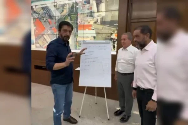 Ricardo Nunes, um homem branco, com camisa azul escura e calça jeans, aponta para um cavalete; outros dois homens, de camisas brancas e calças escuras, estão ao lado direito da imagem - Metrópoles
