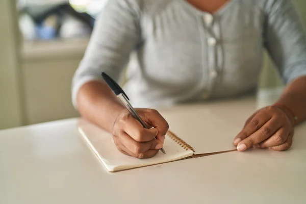 Mulher afro-americana escrevendo em um caderno sentada à mesa em casa. Destra. Metrópoles