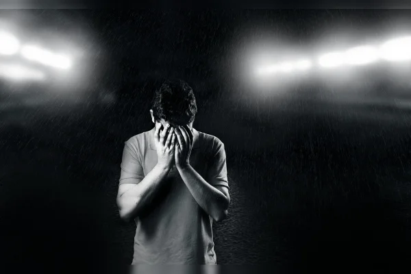 Imagem em preto e branco de um homem com as mãos no rosto, de cabeça baixa, em meio a efeitos que simulam pingos de chuva.