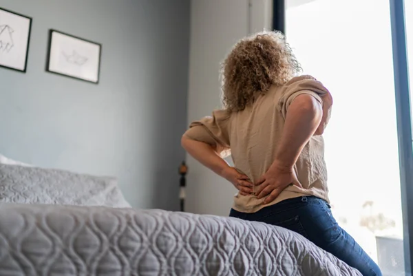 Foto de mulher sentada em cama com mãos nas costas - Metrópoles