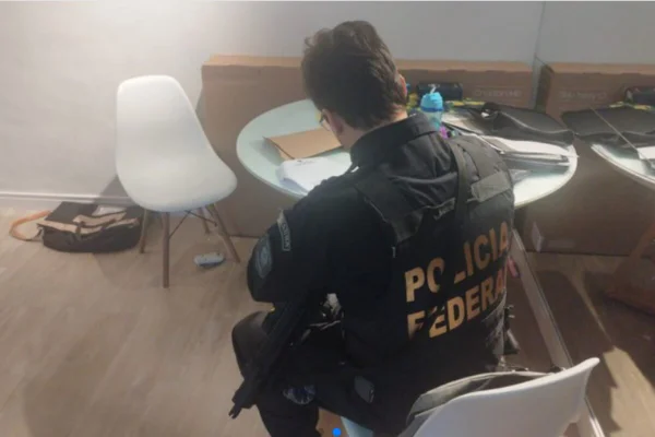 Policial de costas, na qual está escrito Polícia Federal, sentado em cadeira em sala com piso de madeira - Metrópoles