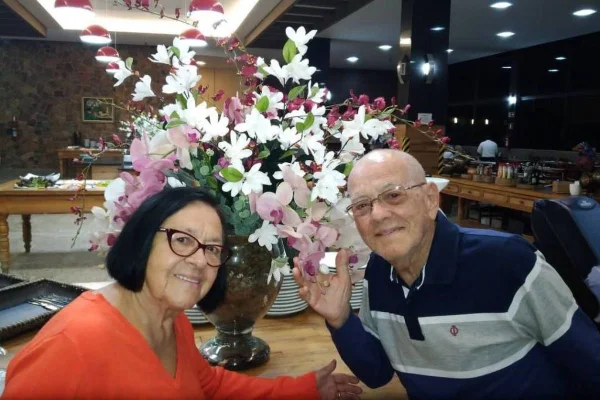 Imagem colorida mostra casal de idosos sorrindo para foto com um arranjo de flores atrás - Metrópoles
