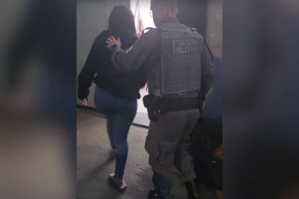 Foto colorida de mulher de costas, sendo conduzida, por policial militar, à delegacia onde foi autuada em flagrante por tentativa de homicídio.