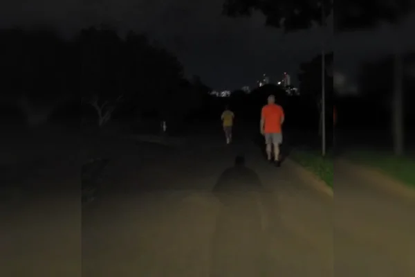 Imagem colorida de uma ciclovia de um parque, com duas pessoas caminhando. O local está escuro devido à falta de iluminação