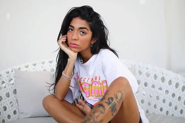 Dhiovanna Barbosa, irmã de Gabigol. Ela está sentada, olhando entediada para a câmera, e tem a pele morena, cabelo longo, liso e preto e usa camisa branca com correntes de prata - Metrópoles