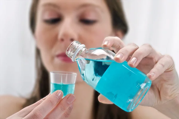 Foto colorida de mulher branca colocando enxaguante bucal dentro de um copo transparente - Metrópoles