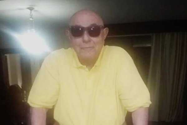 Idoso careca usa óculos escuros e camisa amarela. Ele olha para frente. Foto colorida