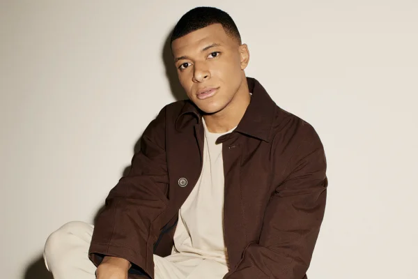 O jogador Kylian Mbappé em foto de campanha da marca de relógios de luxo Hublot. Ele usa uma camiseta branca e, por cima, uma jaqueta marrom. - Metrópoles