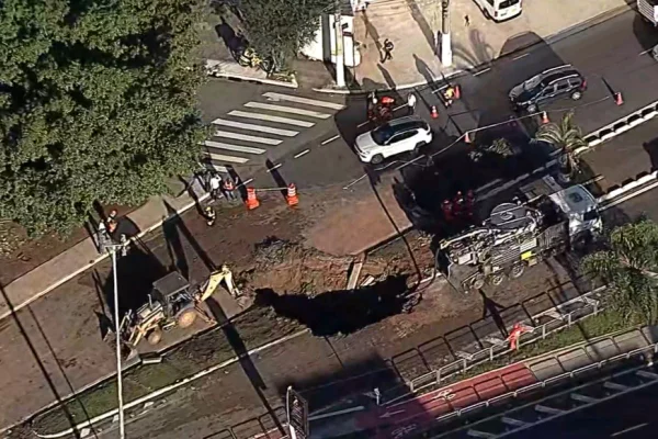 Imagem aérea colorida mostra buraco de grandes dimensões no meio de avenida de concreto. Há uma retorescavadeira ao lado do buraco e um carro passando ao lado - Metrópoles