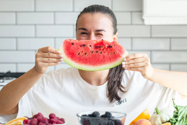 Foto colorida de mulher segurando uma fatia de melancia perdo do rosto, em frente a uma mesa cheia de frutas - Metrópoles.