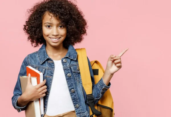 Menina negra de cabelos enrolados com livros nas mãos apontando para o lado com jaqueta jeans - Metrópoles